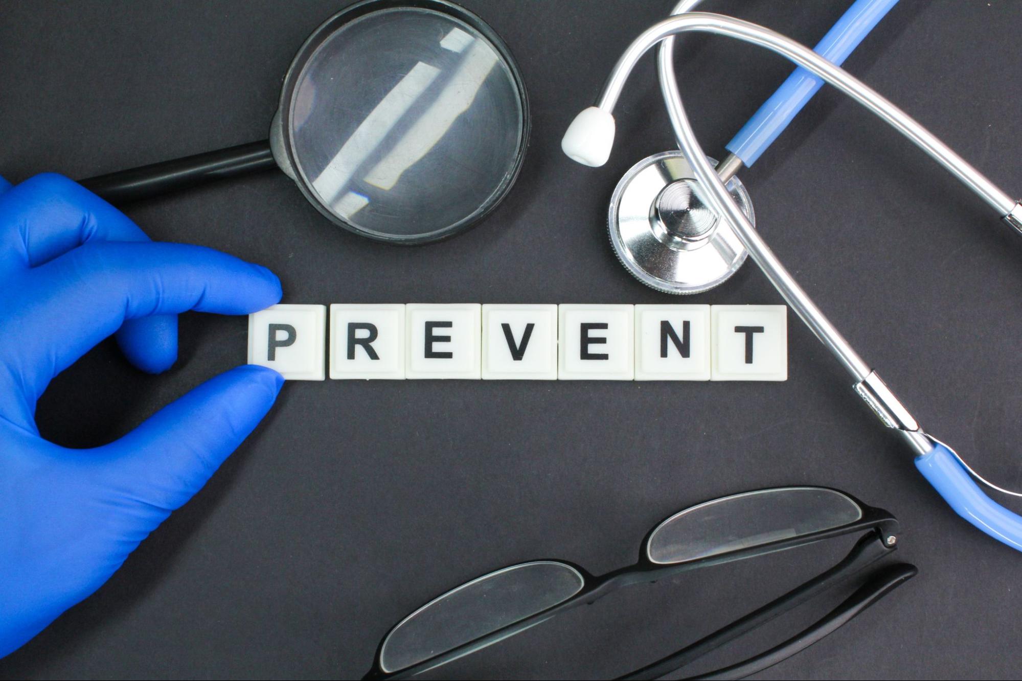 A close-up of someone in a blue latex medical glove spelling out the word “Prevent” in Scrabble-type letters next to a stethoscope, magnifying glass, and glasses.