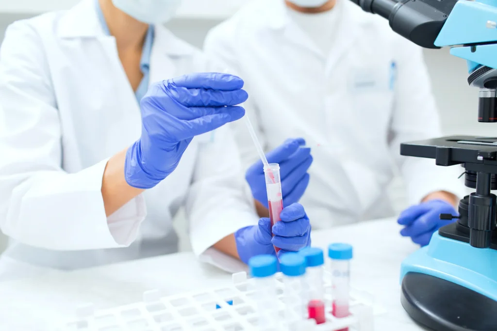 Scientists in lab coats and blue gloves working with a test tube and pipette, with a microscope in the background